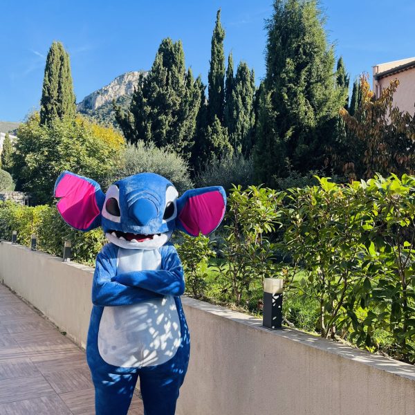 Une mascotte Stitch avec les bras croisés, arborant une expression malicieuse. Le costume est de couleur bleue avec des oreilles larges et des détails fidèles au personnage Disney, capturant son attitude espiègle et confiante.